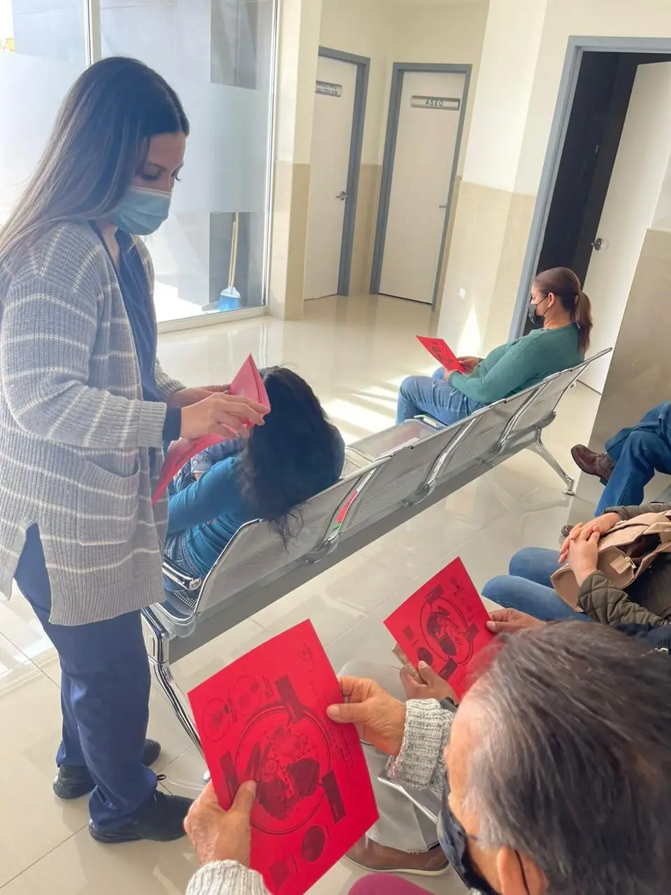 Reyna Isabel Gutierrez Pierce se encuentra en un centro de salud repartiendo hojas con una grafica de comida en el. Las presonas a quienes le esta dando las hojas son cuatro adultos mayores.