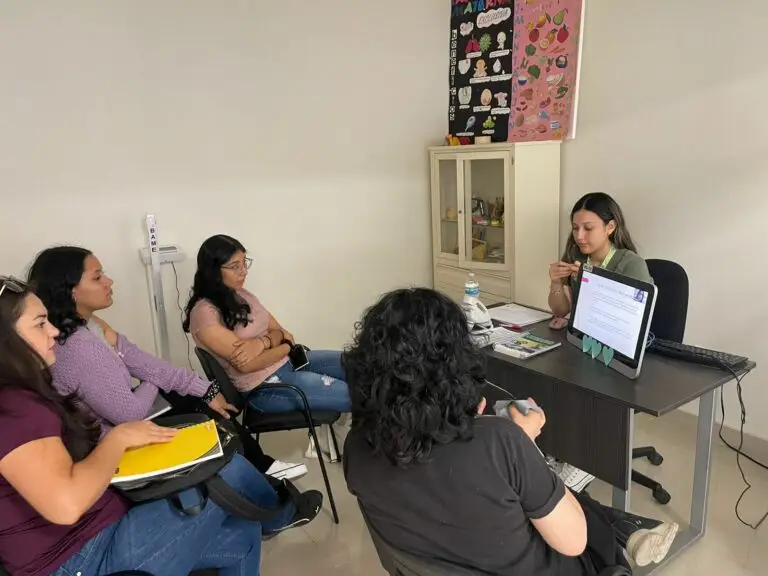 Reyna Isabel Gutierrez Pierce dando una presentacion de zoom a cuatro estudiantes de universidad. Las estudiantes estan sentadas en un semi-circulo, con Reyna enfrente de ellas y un monitor.